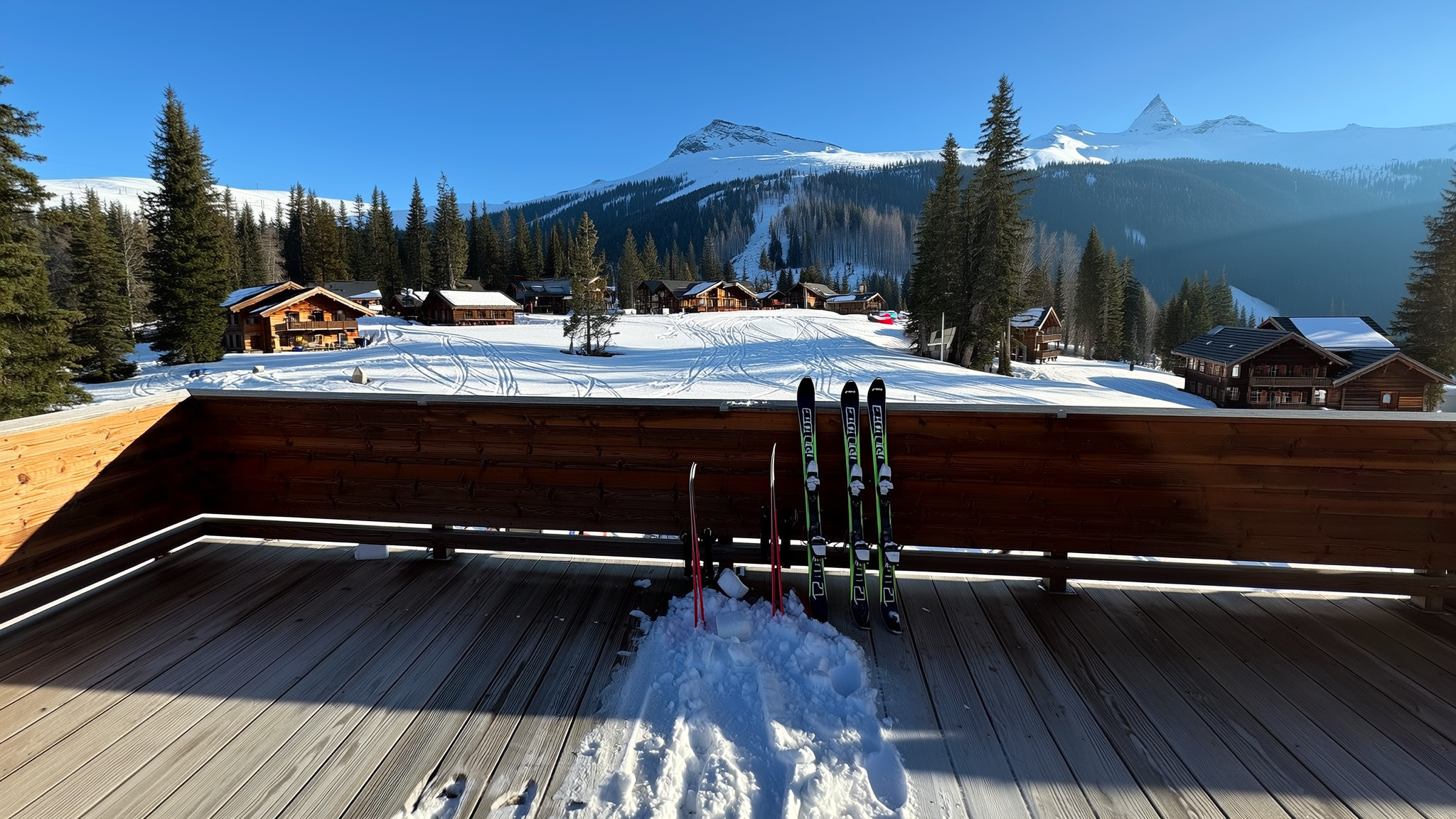 Skis stood upright in the snow on a sunlit timber deck, with lodge buildings and a snow-covered ridge beyond