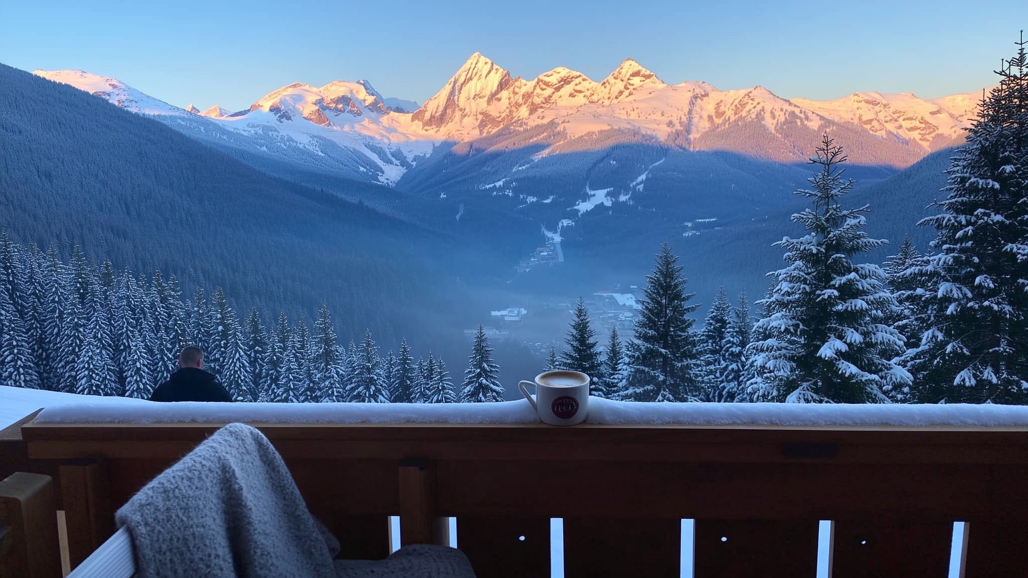 First light on the peaks seen from a snow-covered balcony, a blanket over the chair and a cup on the rail