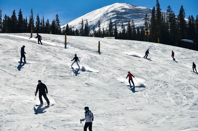 Skiers and snowboarders spread out across a wide groomed run with a bare summit behind