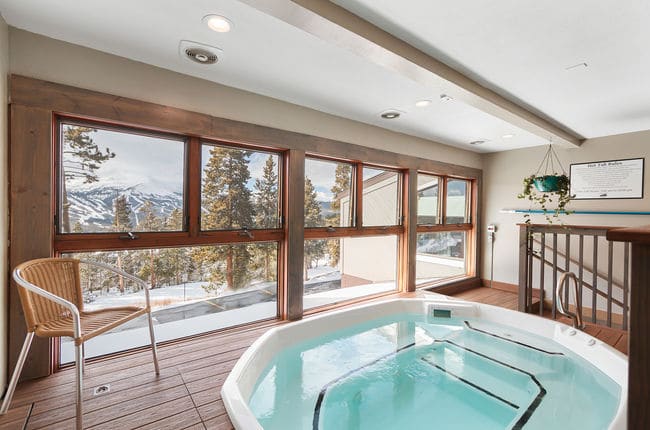 An indoor hot tub with a wall of windows looking out over snow-covered pines and the Tenmile Range